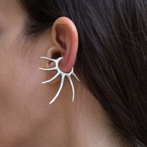 May include: A silver ear cuff with a spiky, sunburst design. The cuff is worn on the upper part of the ear.