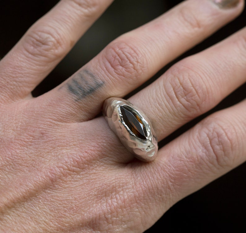 Puede incluir: Un anillo de plata con una piedra preciosa marr&oacute;n engastada en una banda texturizada.