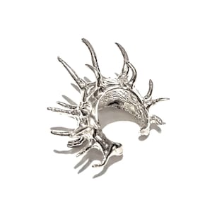 May include: A silver-colored ear cuff with a unique, organic design. The cuff features multiple, irregular, pointed protrusions, creating a textured and sculptural appearance. The jewelry piece is set against a white background.