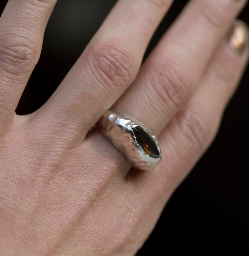 Puede incluir: Un anillo de plata con una gran piedra preciosa ovalada de color marr&oacute;n engastada en una banda texturizada.