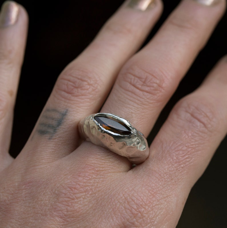 Puede incluir: Un anillo de plata con una banda texturizada y una piedra preciosa ovalada marr&oacute;n.