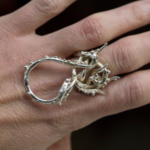 Könnte beinhalten: Ein silberner Ring mit einem einzigartigen, organischen Design. Der Ring besteht aus gedrehtem Metall mit kleinen, runden Perlen an den Rändern.
