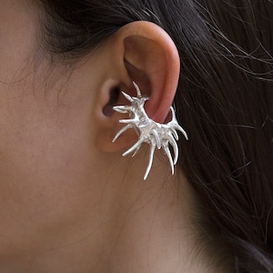 May include: Silver ear cuff with a spiky, organic design. The cuff is worn on the upper part of the ear.