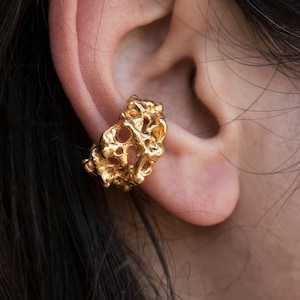 May include: Gold ear cuff with a textured, organic design. The cuff is worn on the upper part of the ear.