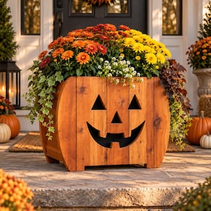 May include: A wooden planter box shaped like a jack-o'-lantern with a carved face featuring triangular eyes and a wide, toothy grin. The natural-toned wood is constructed from vertical planks and is filled with vibrant orange, yellow, and white flowers.