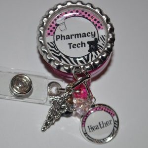 K&ouml;nnte beinhalten: Ein einziehbarer Ausweishalter mit Zebra-Muster in Pink und Schwarz mit einem silbernen Anh&auml;nger in Form eines Caduceus und einem rosa-schwarz gepunkteten Anh&auml;nger mit dem Namen "Heather". Der Ausweishalter hat einen rosa-schwarz gestreiften Zebra-Hintergrund mit dem Text "Pharmacy Tech" und einem Schwarzwei&szlig;bild eines M&ouml;rser und St&ouml;&szlig;els.