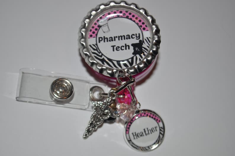 K&ouml;nnte beinhalten: Ein einziehbarer Ausweishalter mit Zebra-Muster in Pink und Schwarz mit einem silbernen Anh&auml;nger in Form eines Caduceus und einem rosa-schwarz gepunkteten Anh&auml;nger mit dem Namen "Heather". Der Ausweishalter hat einen rosa-schwarz gestreiften Zebra-Hintergrund mit dem Text "Pharmacy Tech" und einem Schwarzwei&szlig;bild eines M&ouml;rser und St&ouml;&szlig;els.