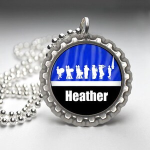 May include: A silver bottle cap necklace with a blue and black design. The design features a silhouette of a marching band and the name "Heather".