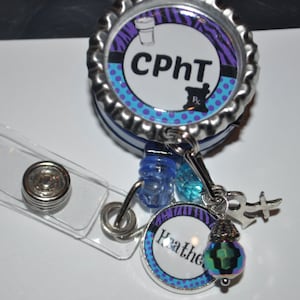 K&ouml;nnte beinhalten: Ein silberner einziehbarer Ausweishalter mit einer blauen, violetten und wei&szlig;en gemusterten Kappe, auf der "CPht" steht, und einem silbernen Anh&auml;nger mit dem Text "Heather".