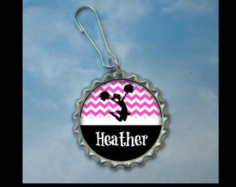 May include: A silver bottle cap keychain with a pink and white chevron pattern, a black silhouette of a cheerleader, and the name "Heather" printed in black.