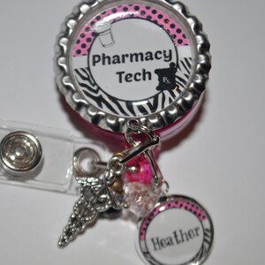 K&ouml;nnte beinhalten: Ein einziehbarer Ausweishalter mit Zebra-Muster in Pink und Schwarz mit einer silbernen Kappe, auf der "Pharmacy Tech" steht, und einem silbernen Anh&auml;nger mit einem Caduceus-Symbol. Der Ausweishalter hat auch einen rosa und wei&szlig;en Kreis mit dem Namen "Heather" darauf.