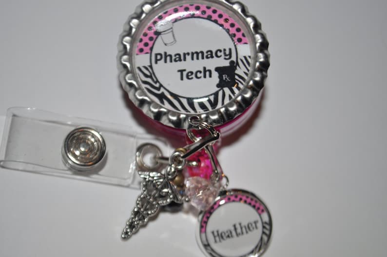K&ouml;nnte beinhalten: Ein einziehbarer Ausweishalter mit Zebra-Muster in Pink und Schwarz mit einer silbernen Kappe, auf der "Pharmacy Tech" steht, und einem silbernen Anh&auml;nger mit einem Caduceus-Symbol. Der Ausweishalter hat auch einen rosa und wei&szlig;en Kreis mit dem Namen "Heather" darauf.