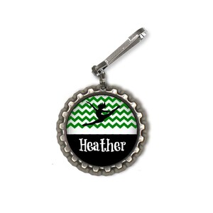 May include: A silver bottle cap keychain with a green and white chevron pattern and a black silhouette of a gymnast. The text "Heather" is printed in white on a black background.