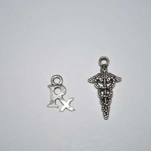K&ouml;nnte beinhalten: Zwei silberne Anh&auml;nger, einer in Form des Buchstabens "R" mit einem Kreuz hindurch, der andere in Form einer Schlange, die sich um einen Stab windet, ein Symbol der Medizin.