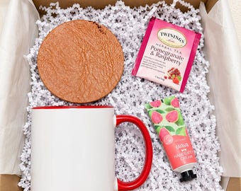 May include: A gift box containing a white ceramic mug with a red handle and interior, a round tan faux leather coaster, a Twinings Pomegranate & Raspberry herbal tea bag, and a tube of guava hand and body cream on white paper filler.