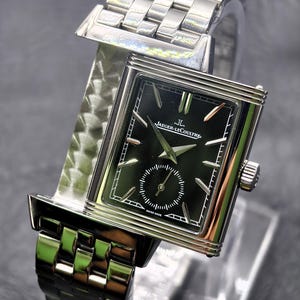 Puede incluir: Un reloj de pulsera rectangular de tono plateado con esfera negra, que presenta el logotipo de Jaeger-LeCoultre, marcadores de hora plateados y un subdial de segundos. El reloj tiene una correa de eslabones de metal pulido y corona.