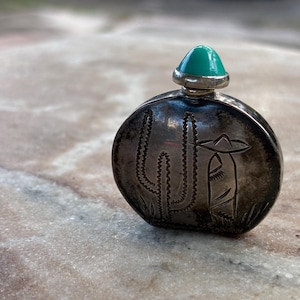 May include: A small, oval-shaped silver bottle featuring an engraved design of a cactus and a figure wearing a wide-brimmed hat. The bottle is topped with a smooth, rounded turquoise stone stopper set in a silver base, creating a compact container.