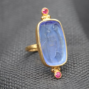 May include: A gold ring featuring a rectangular blue intaglio gemstone carved with a figure. The setting includes a small round pink gemstone at the top and bottom of the central blue stone, all mounted on a textured gold band with a polished finish.