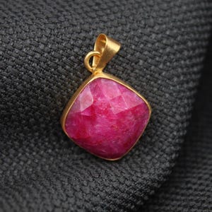 May include: A square-shaped pendant featuring a faceted, vibrant pink gemstone set in a gold-toned metal frame. The pendant includes a small, matching gold-toned bail at the top for attachment to a chain or cord, showcasing a polished finish.