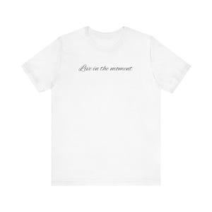 Könnte beinhalten: Ein schlichtes weißes Kurzarm-T-Shirt mit dem schwarzen, kursiven Schriftzug Live in the moment, der mittig auf der Brust aufgedruckt ist. Das Kleidungsstück hat einen klassischen Rundhalsausschnitt und einen geraden Saum.