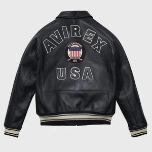Puede incluir: Una chaqueta de cuero negra con el texto AVIREX en letras blancas arqueadas en la parte superior de la espalda. Debajo del texto hay un parche circular con un diseño de bandera estadounidense y las palabras All Americans Established 1975. La palabra USA está impresa en letras blancas grandes en la parte inferior, y la chaqueta tiene puños y dobladillo acanalados con rayas blancas.