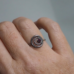 May include: A silver ring with a spiral design and a small, round, red gemstone at the center. The ring is worn on a finger, showcasing its intricate details and the contrast between the metal and the gemstone.