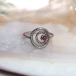 May include: A silver ring featuring a circular design with a textured, dotted surface. The center of the ring contains a small, round, deep pink gemstone set within an inner circle, creating a layered, concentric appearance on a thin band.