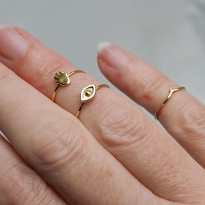 May include: Three gold-toned rings worn on fingers. One ring features a small hamsa hand charm, another displays a central eye-shaped motif, and the third is a simple, thin band with a subtle V-shaped point at the center of the metal wire.