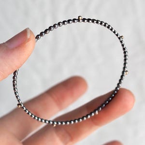 以下が含まれることがあります： 小さな濃いグレーのビーズで構成された繊細なブレスレット。ブレスレットは、小さな金色のビーズでアクセントをつけ、微妙なコントラストを生み出しています。ジュエリーは手で持たれており、その円形デザインと繊細な職人技が示されています。