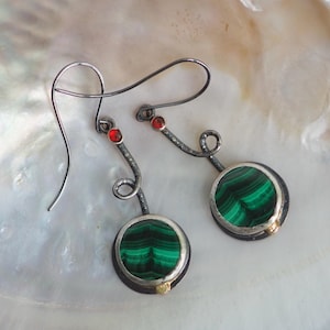 Könnte beinhalten: Ein Paar Ohrhänger mit runden Malachit-Cabochons in dunkel oxidiertem Metall. Jeder Ohrring hat einen geschwungenen, texturierten Stiel mit einem kleinen roten Steinakzent oben und einem goldfarbenen Detail an der Basis der Fassung.