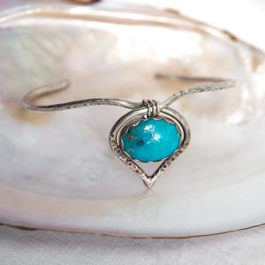 May include: A silver cuff bracelet featuring a central teardrop-shaped frame with a hammered texture. The frame holds a smooth, oval-shaped turquoise stone with dark matrix inclusions. The silver band is secured to the frame with wire wrapping.