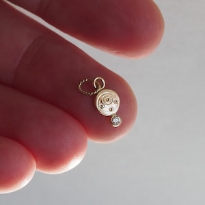 May include: A small, gold-coloured pendant with a circular design and a small, clear gemstone. The pendant is held between fingers, showcasing its delicate size and intricate details. The pendant's design includes concentric circles and a small, sparkling stone.