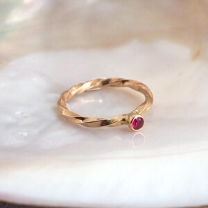 May include: A gold ring featuring a twisted band design and a single, small, round, vibrant pinkish-red gemstone set in a bezel setting. The metal has a polished finish, and the band exhibits a continuous spiral or rope-like texture throughout.
