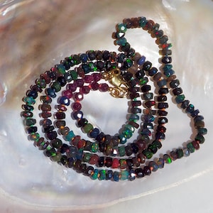 May include: A necklace composed of small, faceted, dark-toned opal beads displaying iridescent flashes of green, blue, and red. The strand features a gold-colored metal lobster clasp and is arranged in a coiled, circular pattern on a smooth surface.
