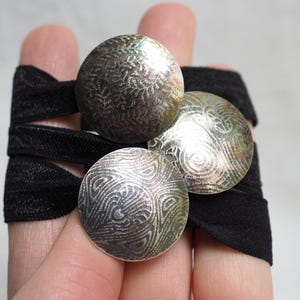 May include: Three circular silver-toned metal discs with intricate etched patterns attached to a black elastic band. Each disc features a unique, detailed texture with a metallic sheen, arranged in a cluster on the dark fabric strap.
