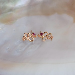 May include: A pair of gold stud earrings shaped like coiled snakes, each featuring a small, round, deep red gemstone set into the head of the snake. The earrings have a polished metallic finish and a delicate, winding design.