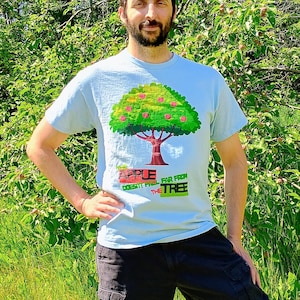 Puede incluir: Una camiseta de manga corta de color azul claro con un gráfico de un manzano verde con frutos rojos. El texto impreso en la camiseta dice "APPLE DOESN'T FALL FAR FROM THE TREE" en letras rojas, verdes y negras en el centro del pecho.