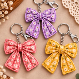 May include: Three fabric bow keychains featuring a repeating floral pattern with small white daisies. Each bow is attached to a silver-toned metal split ring and a lobster clasp. The bows are colored in purple, pink, and yellow respectively.