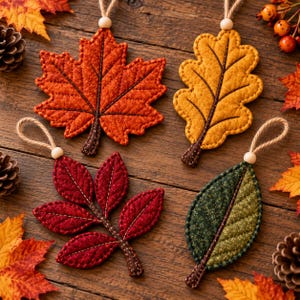 May include: Four fabric leaf ornaments with stitched details and hanging loops. The set includes a burnt orange maple leaf, a golden yellow oak leaf, a deep red compound leaf, and a dark green oval leaf, each with a brown stem.