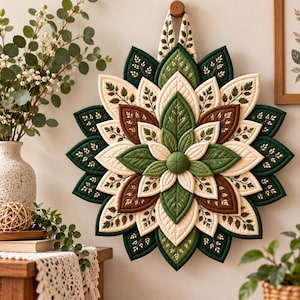 Op de afbeelding: Een ronde stoffen wandhanger met een gelaagd, driedimensionaal bloemendesign. De bloemblaadjes zijn gemaakt van doorgestikte stof in tinten donkergroen, crème en bruin, met geborduurde bladpatronen op de crèmekleurige delen.