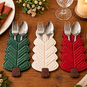 May include: Three quilted fabric utensil holders shaped like pine trees in dark green, cream, and red. Each holder features a brown rectangular base and contains a silver metal fork and spoon. The quilted texture follows a leaf-like pattern.
