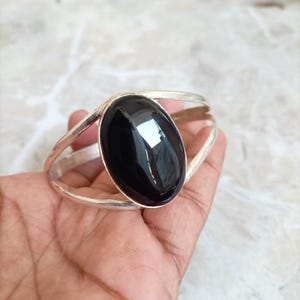 Peut inclure: Un bracelet manchette en argent orné d'une grande pierre noire ovale et lisse fixée au centre. La bande métallique se divise en deux bras distincts qui se courbent autour du poignet, offrant une finition polie et réfléchissante au bijou.