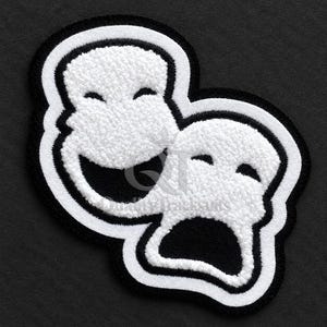 May include: A black and white embroidered patch featuring two theatrical masks, one smiling and one frowning, with a textured chenille-style finish and a thick border. The text Quality Tracksuits is visible in the center of the design.