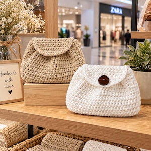 May include: Two small crochet pouches with flap closures. One pouch is beige and the other is white, featuring a single dark brown circular button on the front flap. The pouches have a textured, woven appearance and are displayed on a wooden shelf.