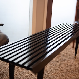 May include: A black wooden bench with a slatted top and black metal legs. The bench is sitting on a woven rug.
