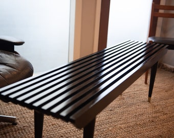 May include: A black wooden bench with a slatted top and black metal legs. The bench is sitting on a woven rug.