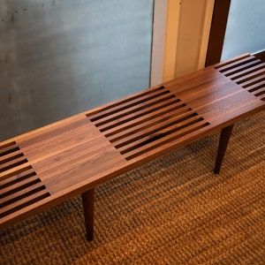 May include: A long, wooden bench with a slatted design. The bench is made of dark wood and has a natural finish. It is sitting on a woven rug.