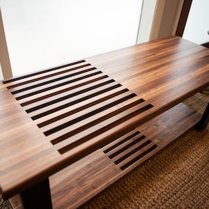 May include: A wooden bench with a slatted design on the top and a lower shelf. The bench is made of dark wood and has a natural finish.