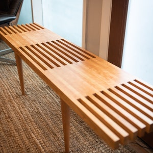 May include: A long, wooden bench with a slatted design. The bench is made of light-colored wood and has a natural finish. The bench is sitting on a woven rug.