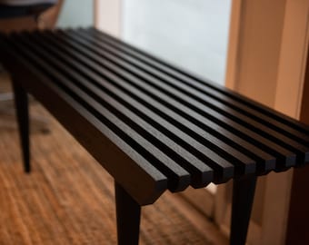 May include: A black wooden bench with a slatted top and black legs. The bench is made of solid wood and has a simple, modern design.